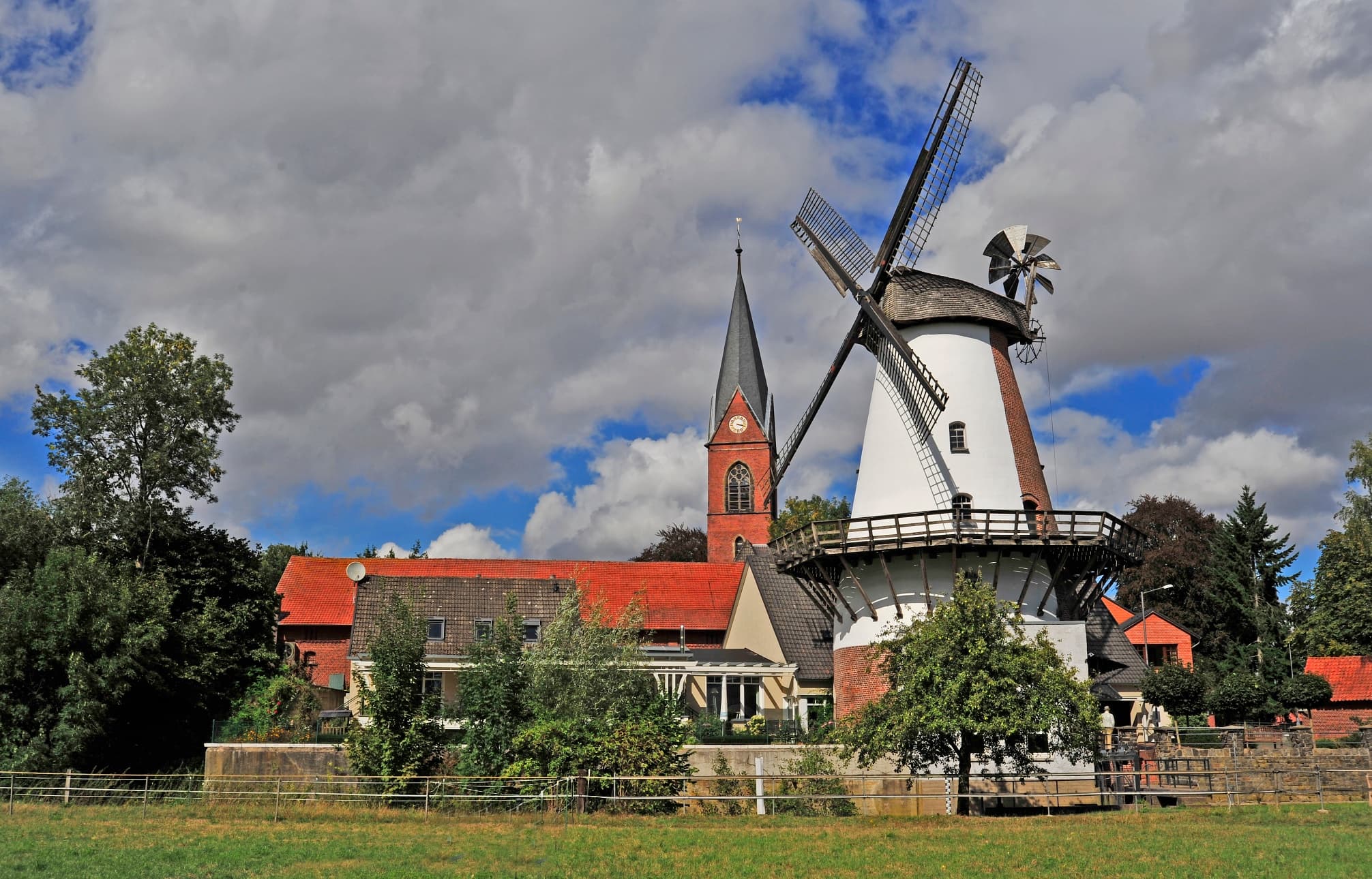 Historische Windmühle neben einer Kirche, umgeben von Bäumen und einem leichten Wolkenhimmel.