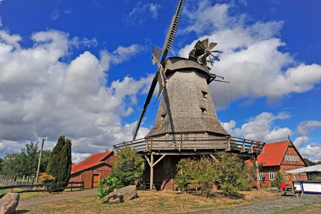 Historische Windmühle mit grauen Schindeln, Holzgalerie und Flügeln vor blauem Himmel und Wolken.
